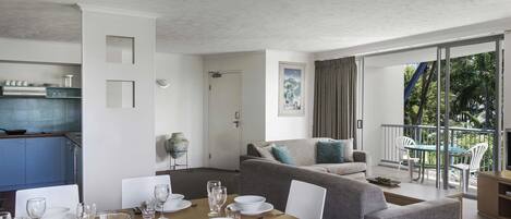 A modern living room with a dining area, a kitchen, and a balcony with a view of greenery.