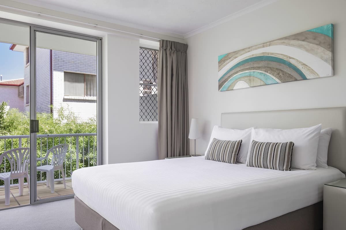 A hotel room with a bed, a nightstand, a painting, a window with curtains, and a view of the outside.