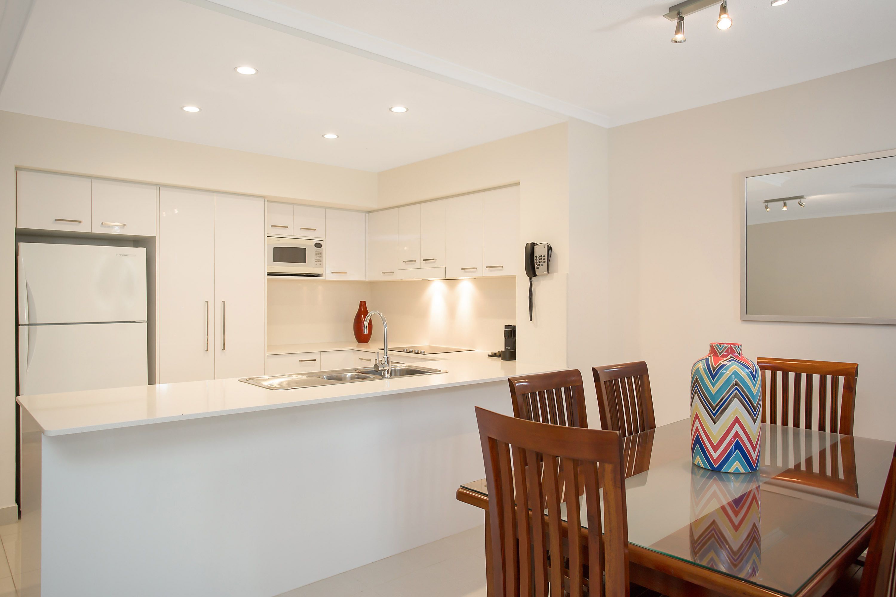 A modern kitchen with white cabinetry, a built-in oven, and a large island with a sink. There are wooden chairs around a glass table and a colorful vase on the island.
