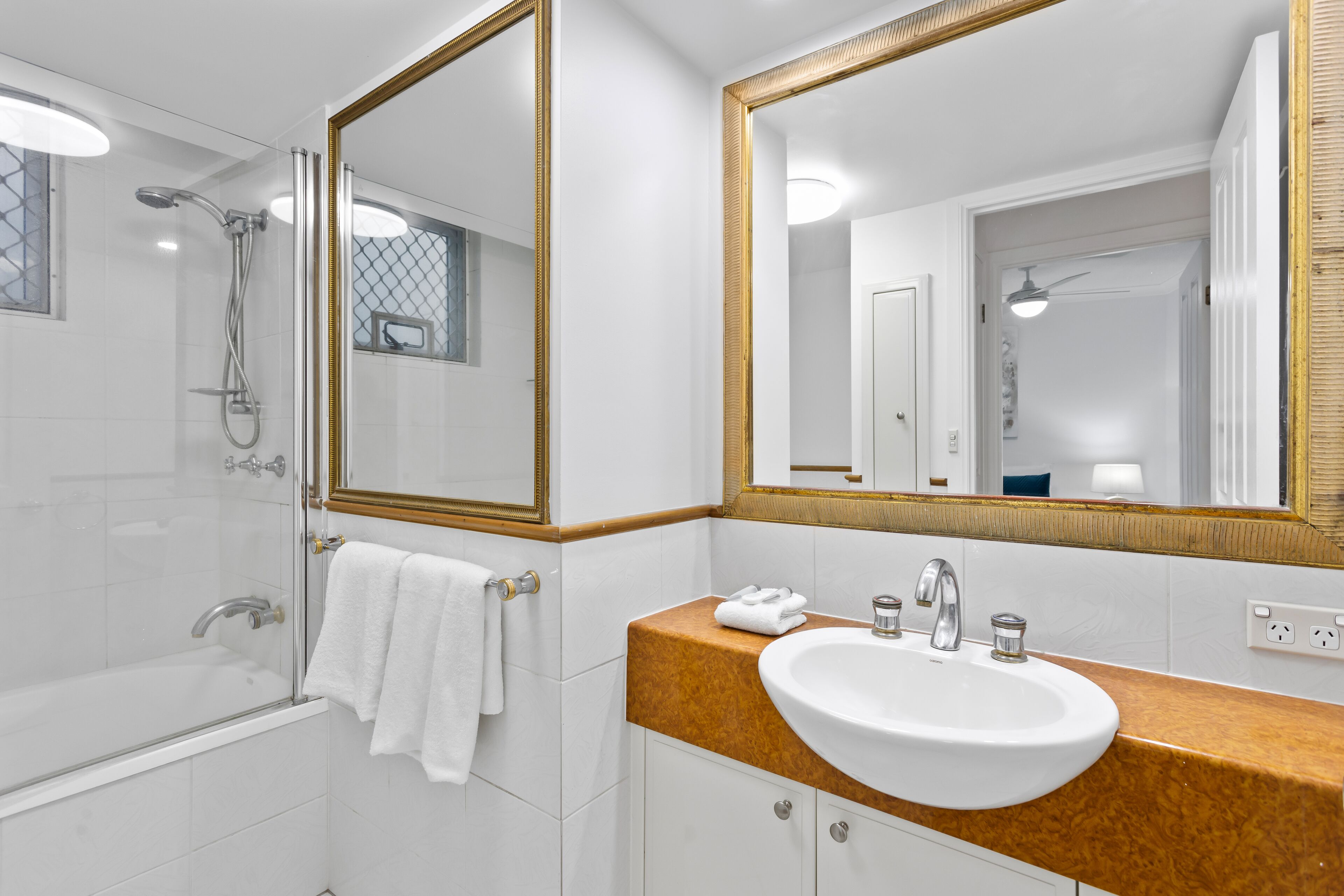 A modern bathroom with a glass-enclosed shower, a white sink with a marble countertop, and a large mirror with a gold frame.