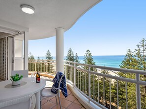 Un balcon avec vue sur l’océan, une table avec une bouteille et des verres, et une chaise avec une serviette.