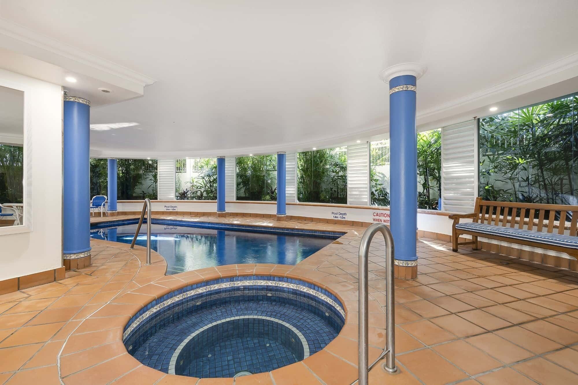 An indoor swimming pool with a hot tub, surrounded by blue columns and a bench.