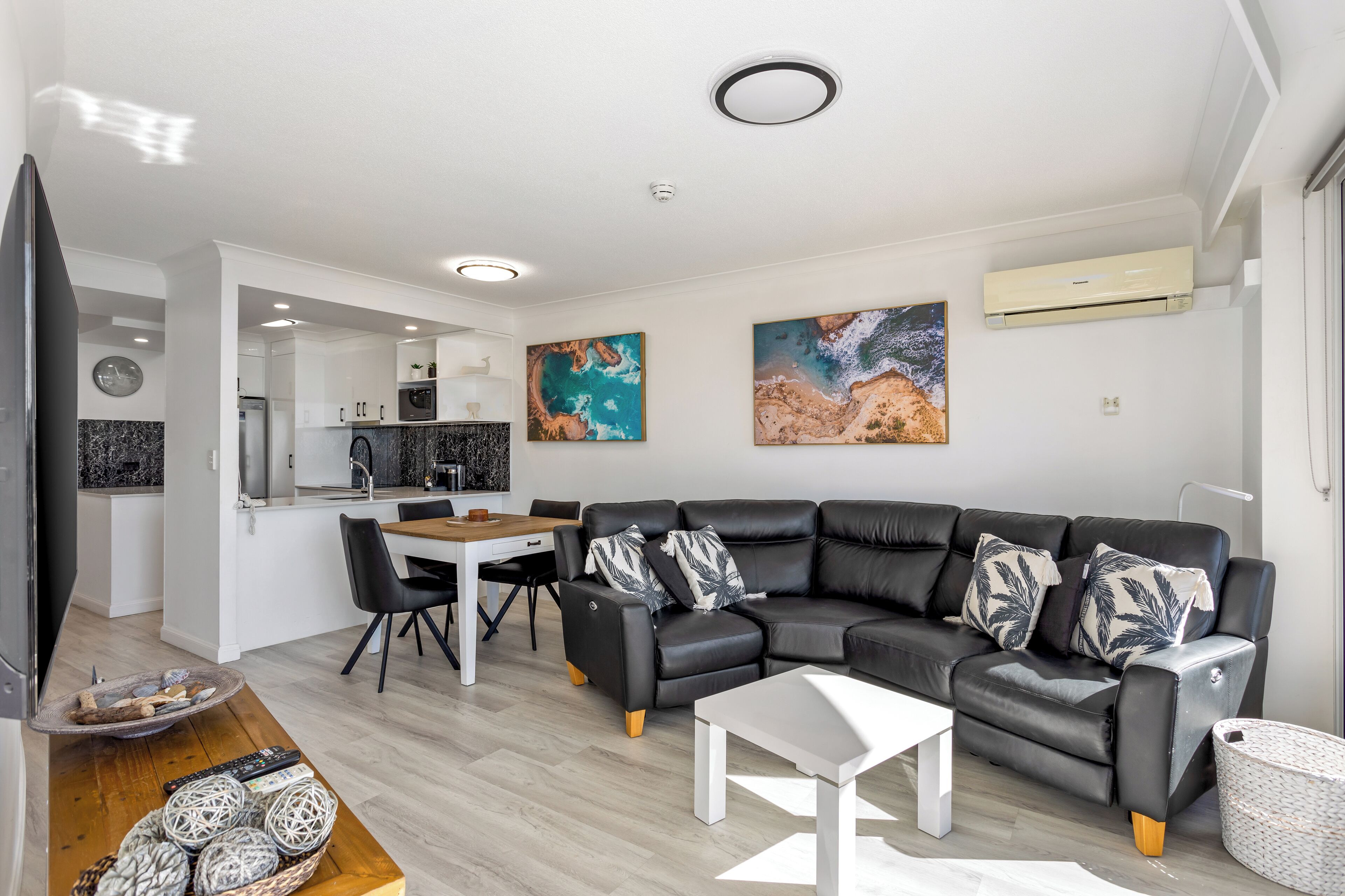 A modern living room with a black leather sofa, a dining table, and a kitchen in the background.