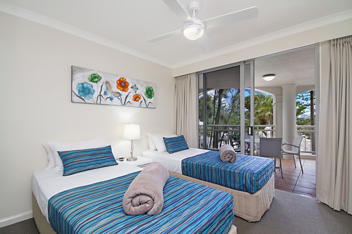 Habitación de hotel con dos camas, ventilador de techo, pintura de flores y balcón con vistas a palmeras.