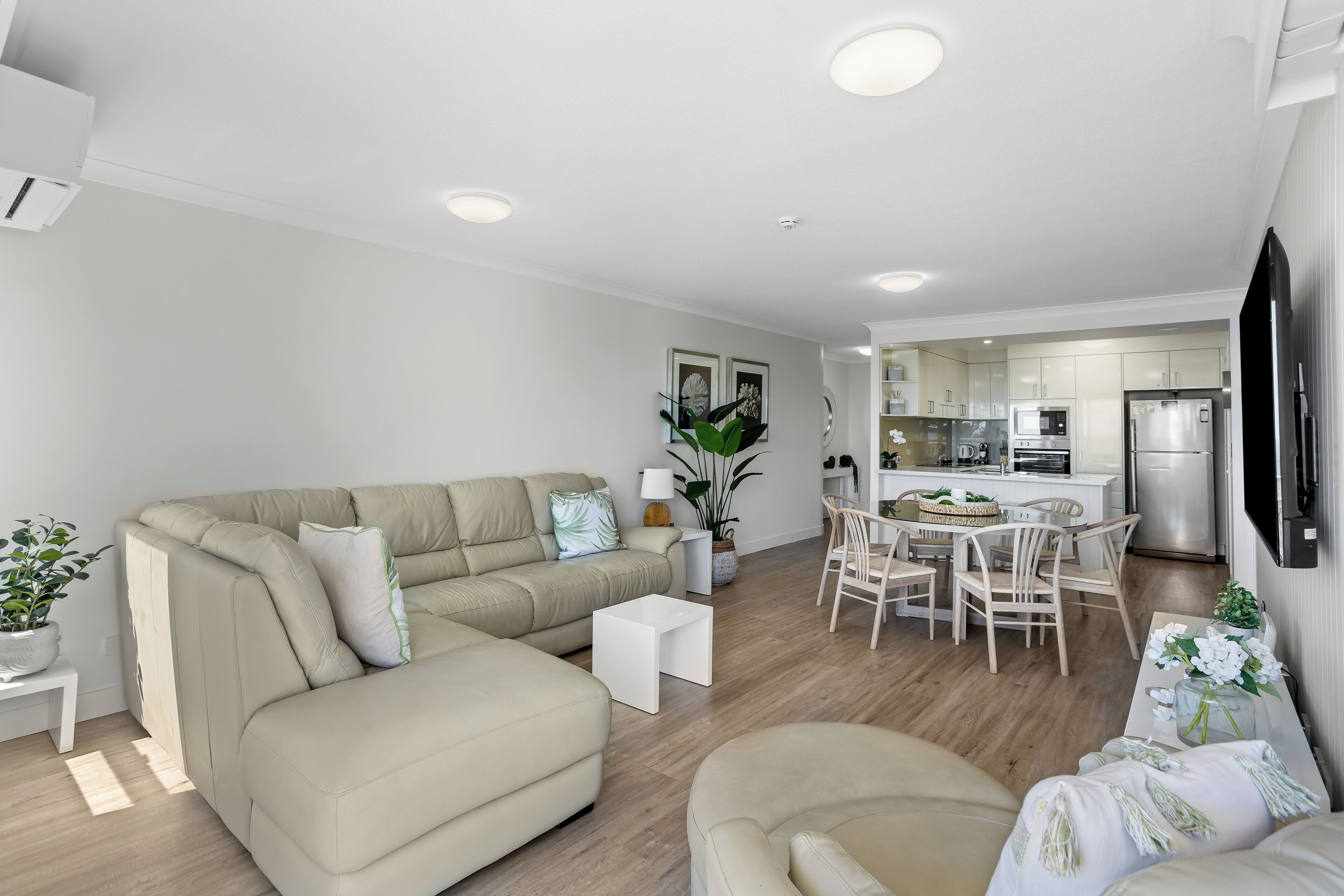 A modern living room with a beige sectional sofa, a dining area with a white table and chairs, and a kitchen with white cabinets and stainless steel appliances.