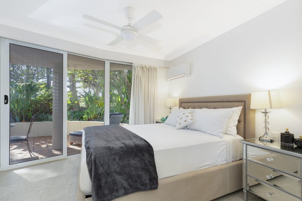 Un dormitorio con cama, mesita de noche, ventilador de techo, ventana con vistas a la vegetación y puerta corrediza de vidrio que da a un espacio exterior.