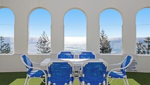 Una habitación con una mesa y cuatro sillas azules, ventanas arqueadas grandes y vista al mar.