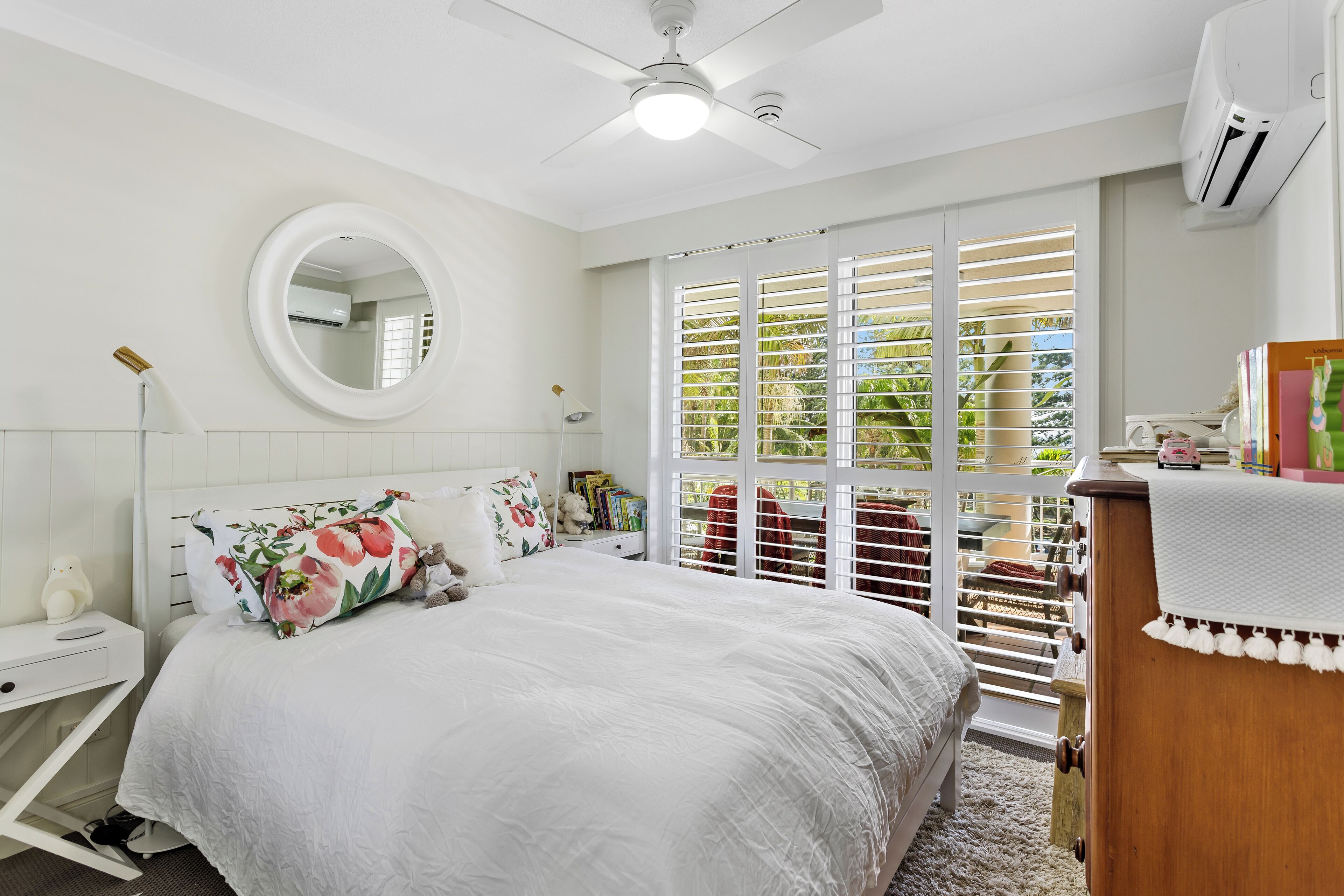 A bedroom with a bed, a round mirror, a ceiling fan, and a window with blinds.