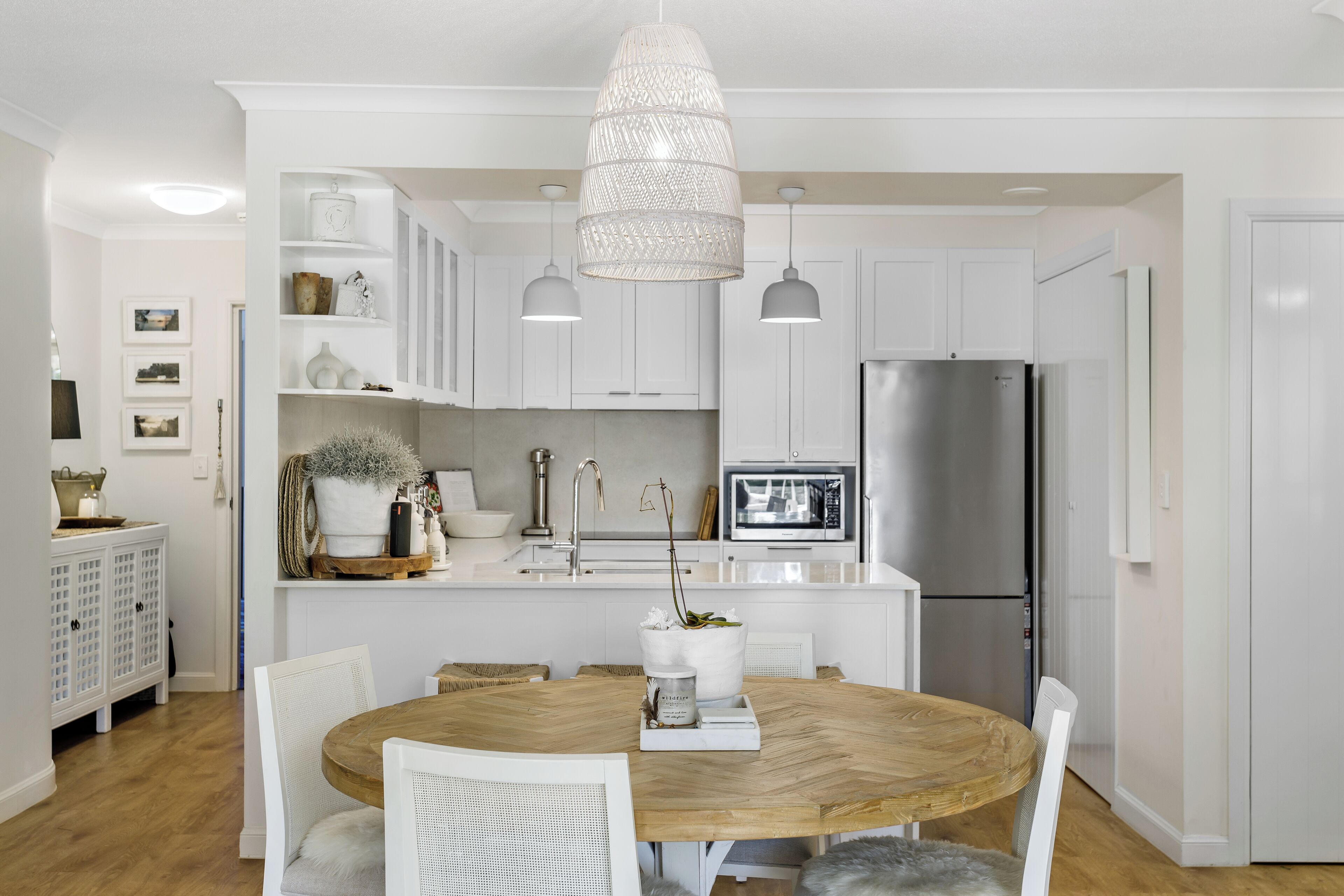 A modern kitchen with white cabinetry, a central island with a wooden table, white chairs, pendant lights, and a stainless steel refrigerator.