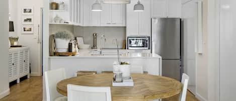 A modern kitchen with white cabinetry, a central island with a wooden table, white chairs, pendant lights, and a stainless steel refrigerator.