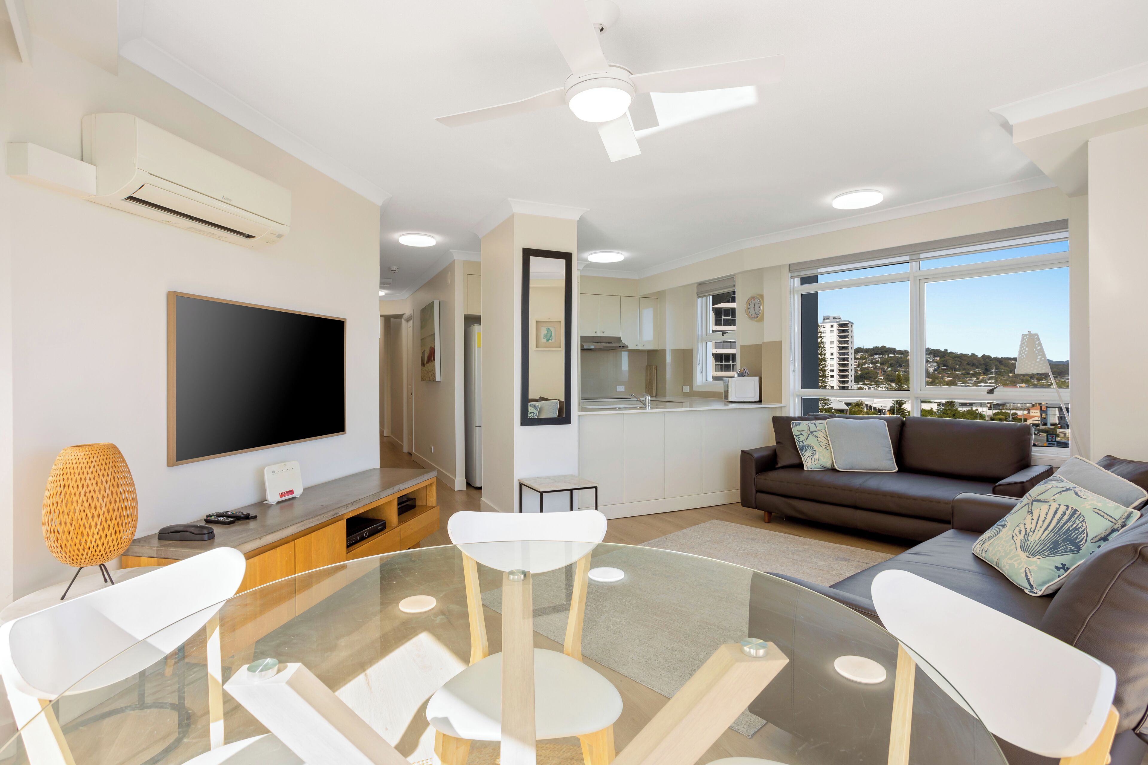 A modern living room with a flat-screen TV, a glass dining table, a sofa, and a city view through large windows.
