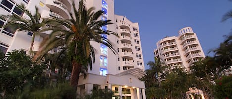 A multi-story hotel with balconies, palm trees, and a well-lit entrance.