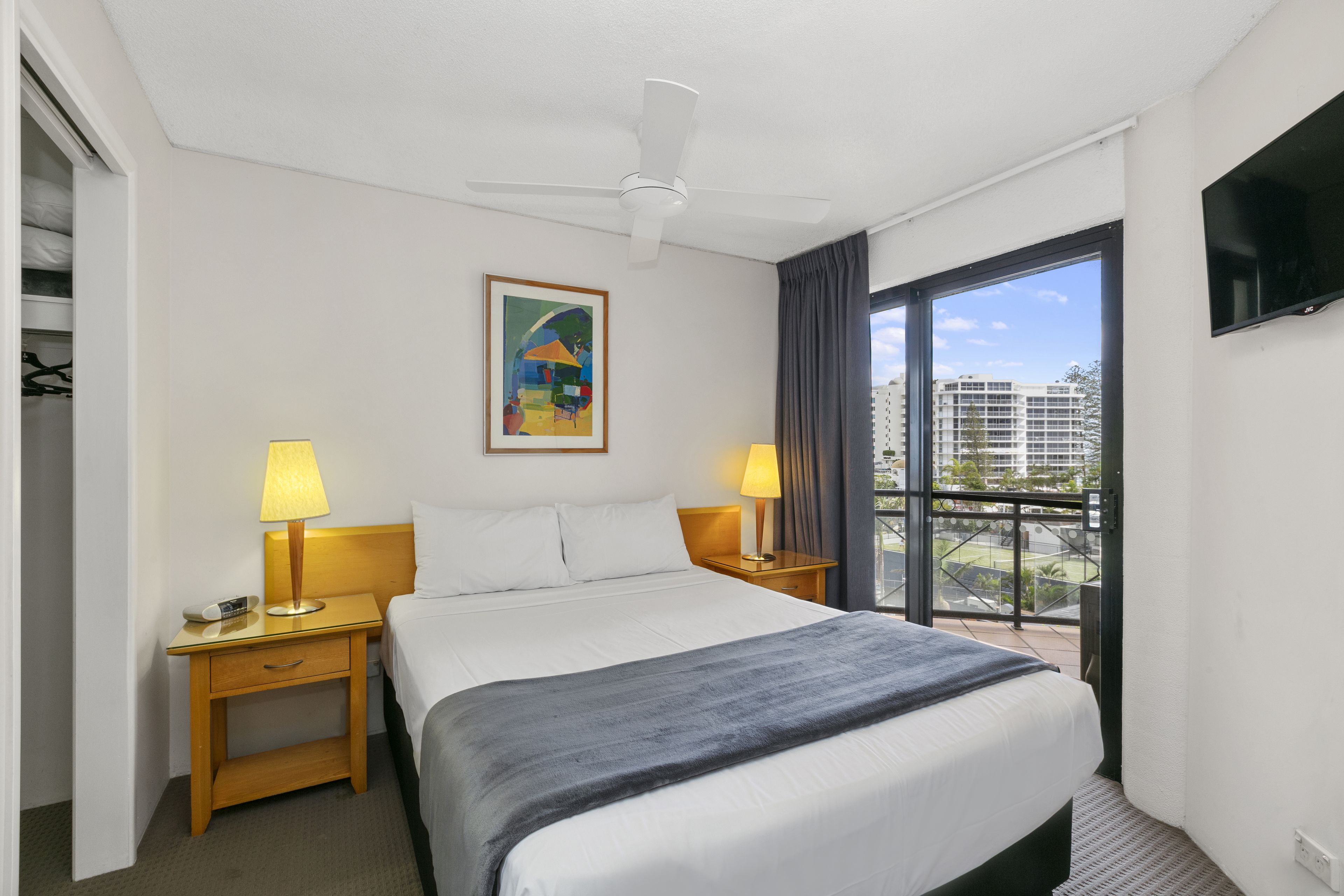 A hotel room with a bed, two bedside tables with lamps, a ceiling fan, a painting on the wall, a TV mounted on the wall, and a balcony with a view of buildings.