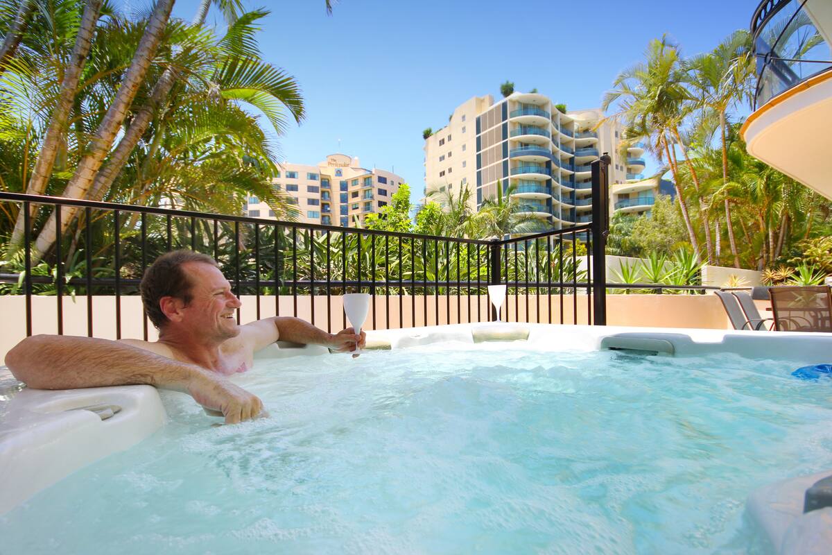 A man relaxing in a hot tub with a view of palm trees and hotel buildings in the background.