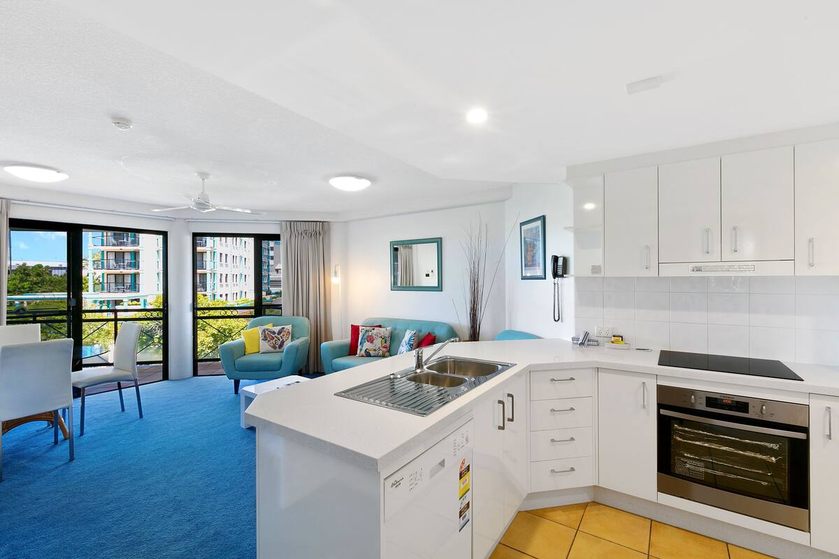 A modern kitchen with white cabinets, a built-in oven, and a sink. There is a blue carpeted area with a sofa and chairs.