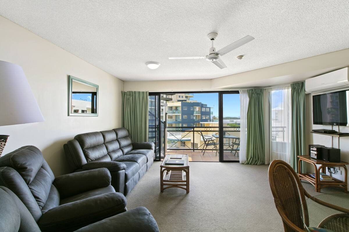 A spacious living room with a leather sofa, a small wooden table, and a wicker chair. A ceiling fan and a flat-screen TV are visible. The room has large windows with green curtains, offering a view of buildings and a balcony outside.
