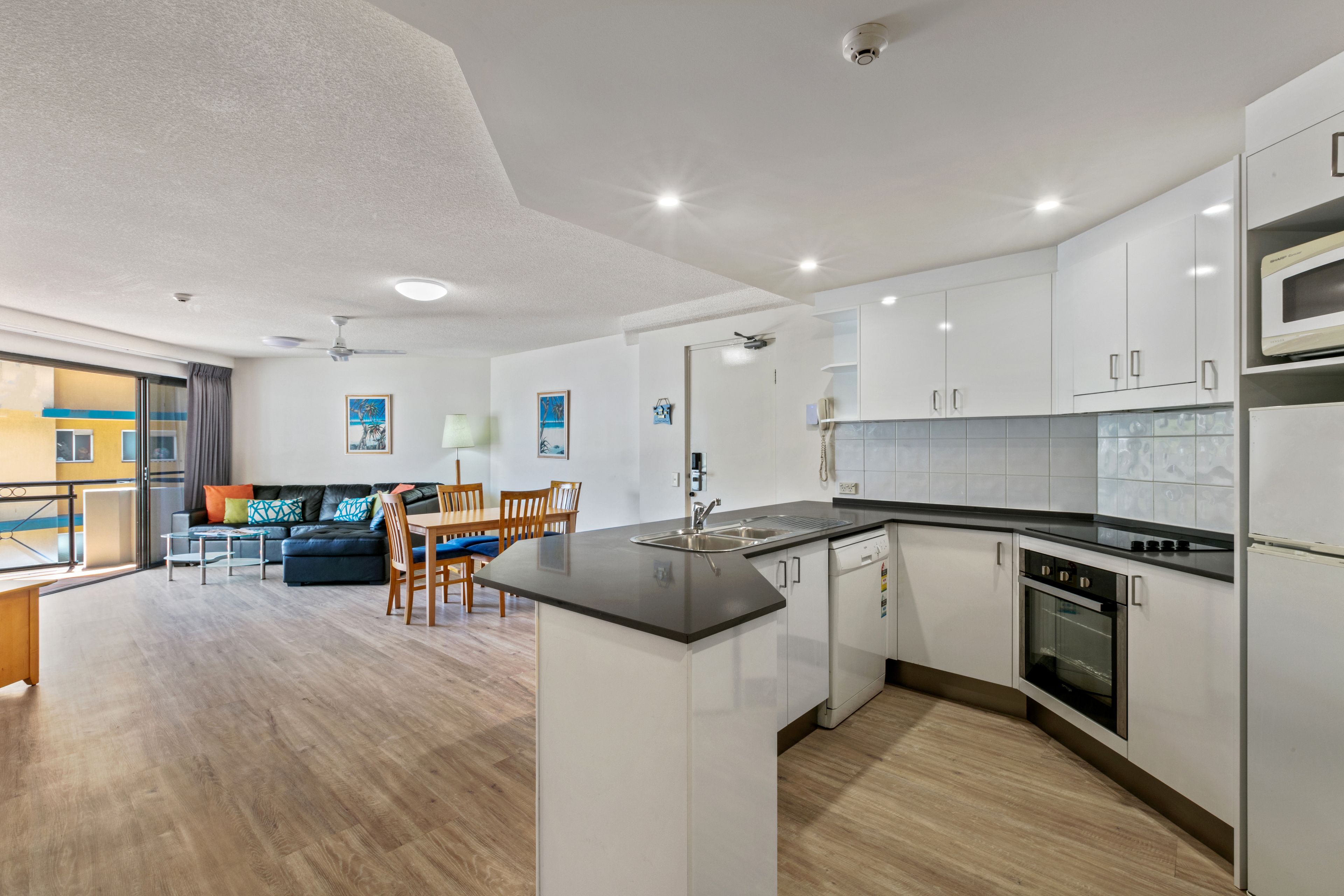 A modern kitchen with white cabinets, a black countertop, and a built-in oven. There is a dining area with a table and chairs, and a sofa in the background.