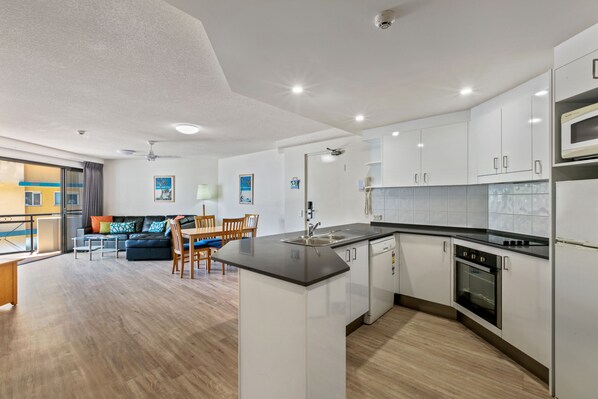A modern kitchen with white cabinets, a black countertop, and a built-in oven. There is a dining area with a table and chairs, and a sofa in the background.