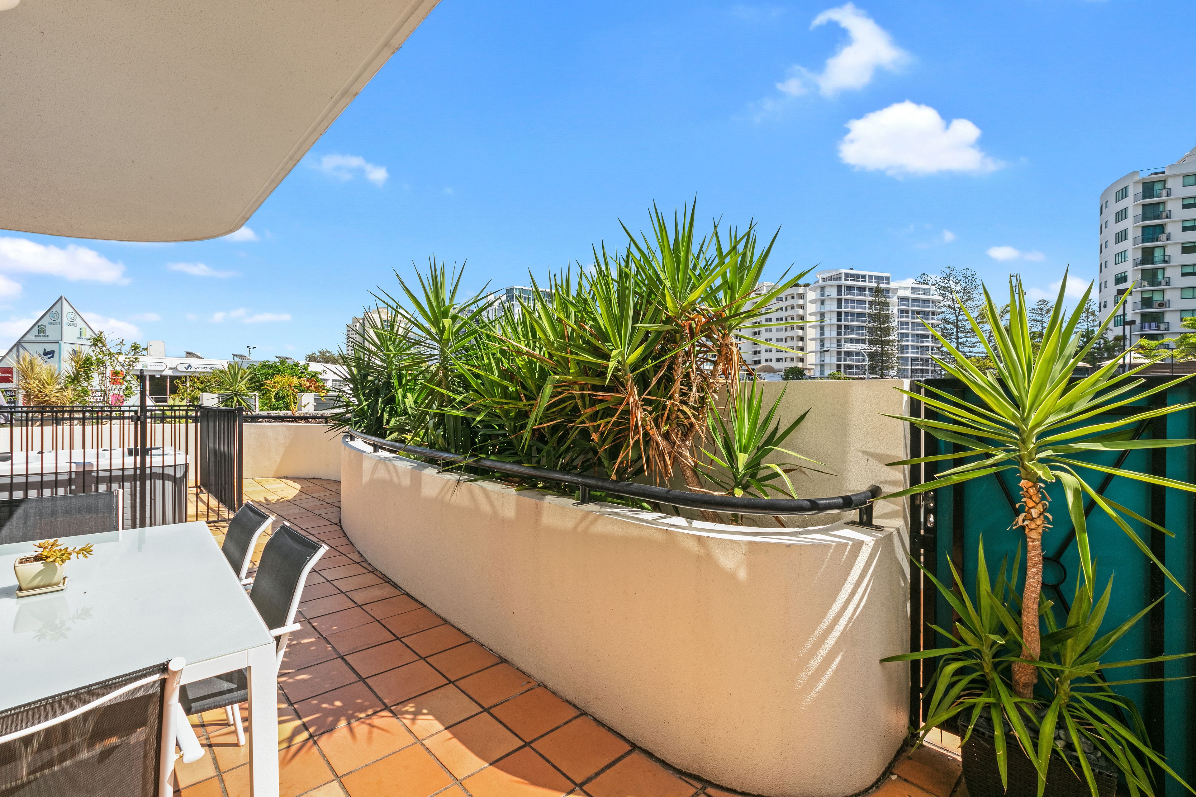 A rooftop terrace with a white table and chairs, surrounded by potted plants and a view of city buildings.