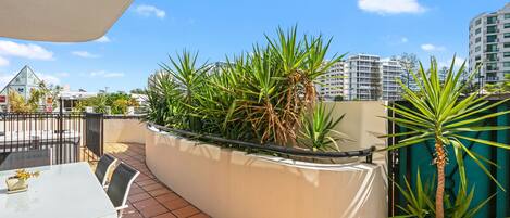 A rooftop terrace with a white table and chairs, surrounded by potted plants and a view of city buildings.