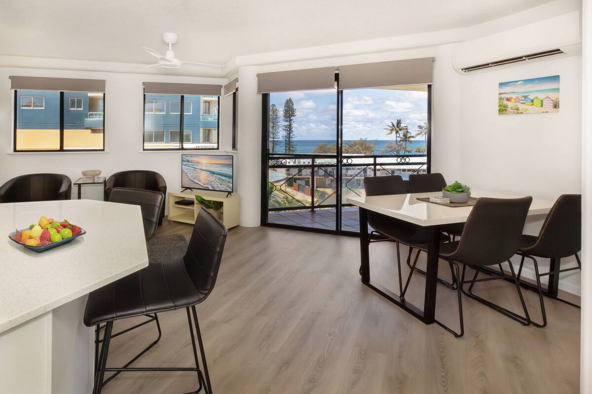 A modern kitchen with a dining area, large windows with a view of the sea, and a balcony with a railing.