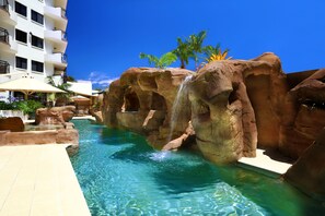 Zona de piscina con cascada, palmeras y un edificio con balcones.