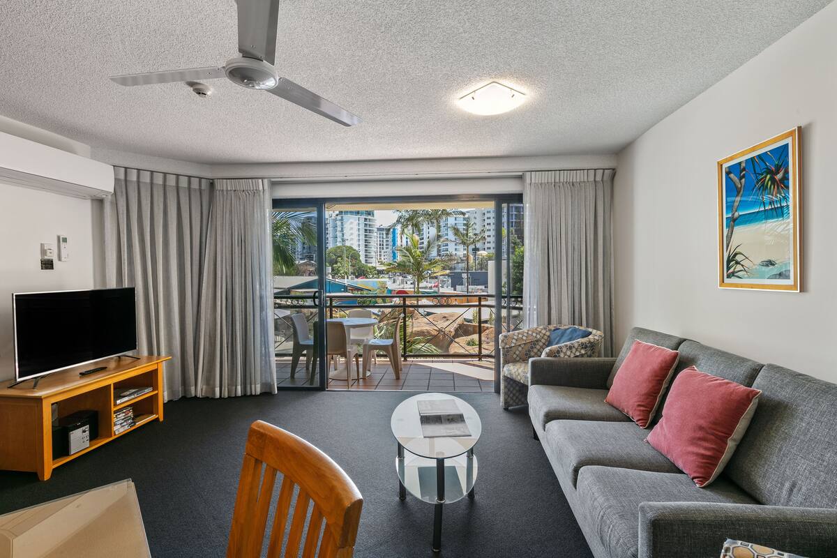 A modern living room with a flat-screen TV, a coffee table, a sofa, and a balcony with a view of palm trees and buildings.