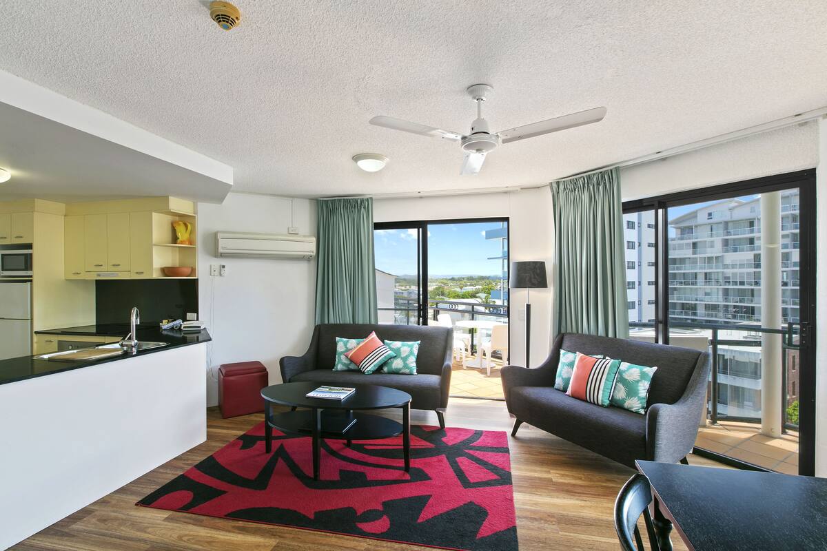 A modern living room with a kitchen area, a ceiling fan, and a large window offering a city view.