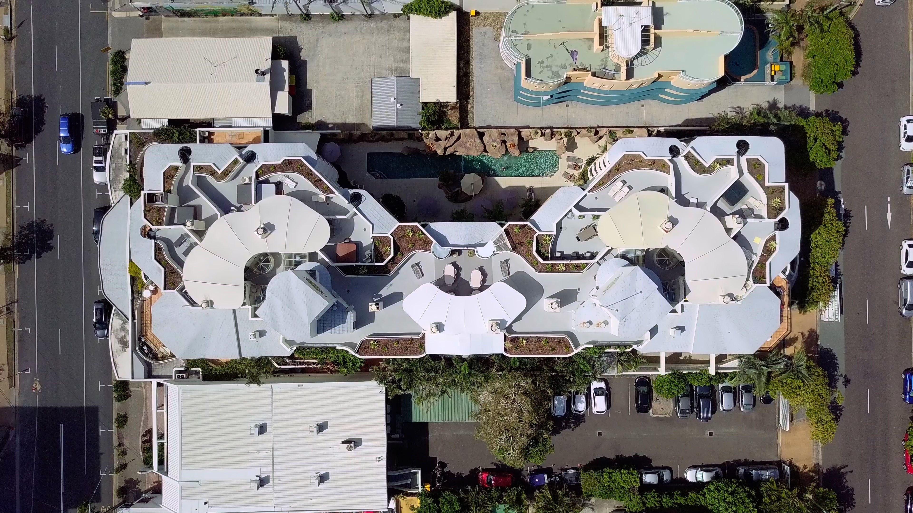 An aerial view of a modern building with a unique roof design, a swimming pool, and a parking lot.