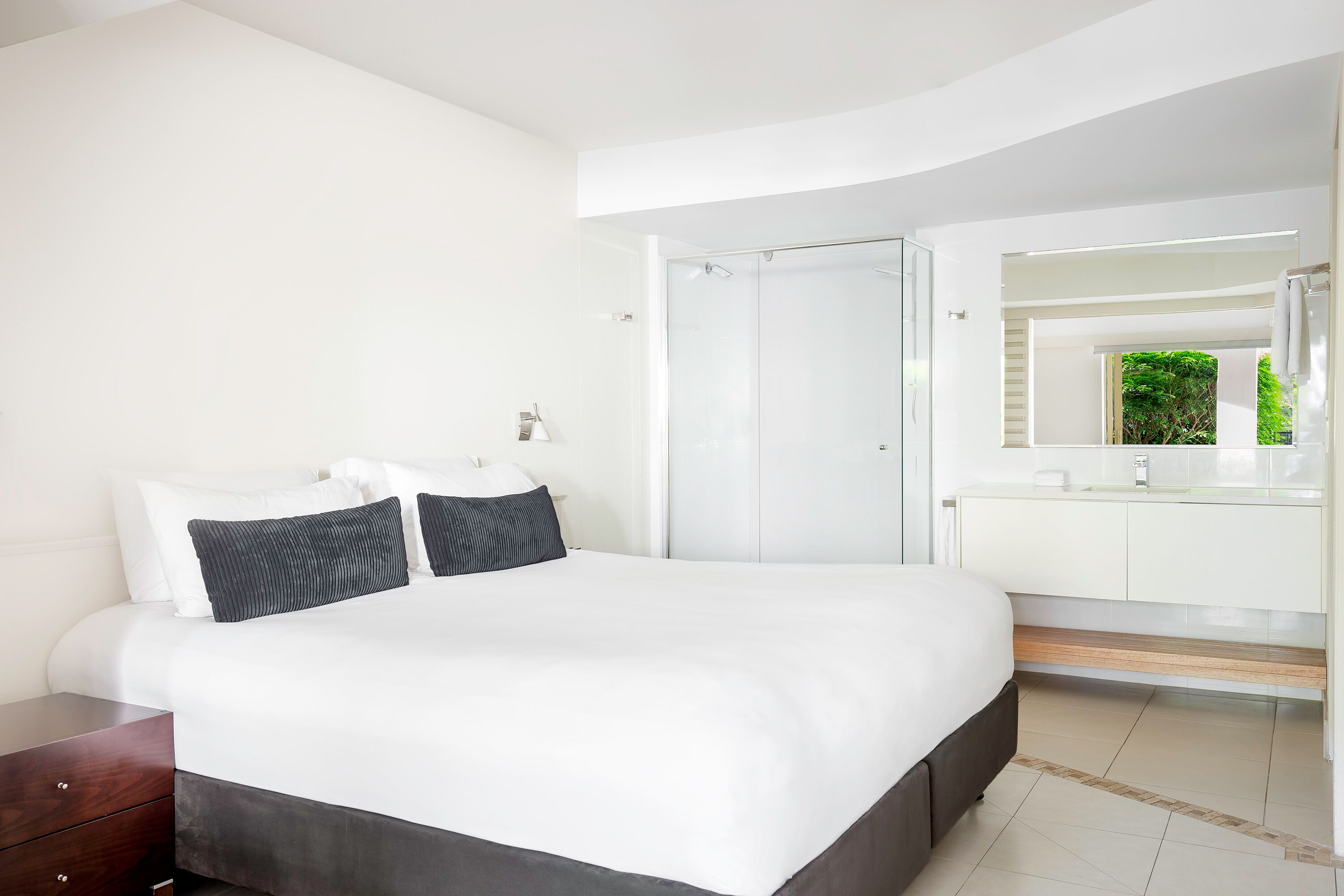 Une chambre moderne avec un grand lit, une table de chevet en bois, une salle de bain blanche dotée d’une douche en verre, et une vue sur la verdure à l’extérieur.