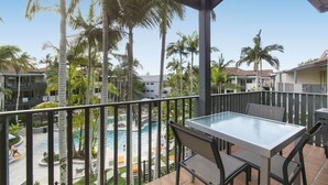 Ein Balkon mit Glastisch und Stühlen, der einen Blick auf ein Schwimmbecken und Palmen bietet.