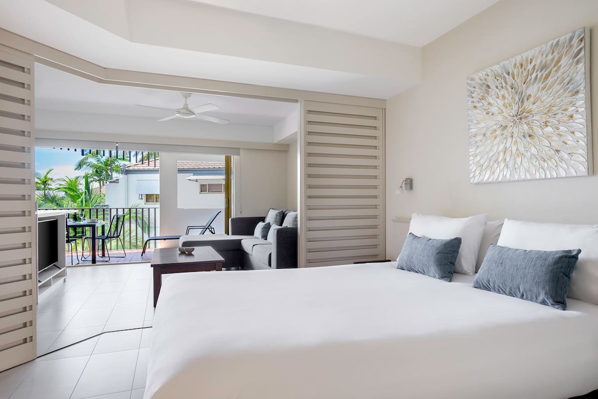 Een moderne hotelkamer met een groot bed, een balkon met een tafel en stoelen, en uitzicht op palmbomen.