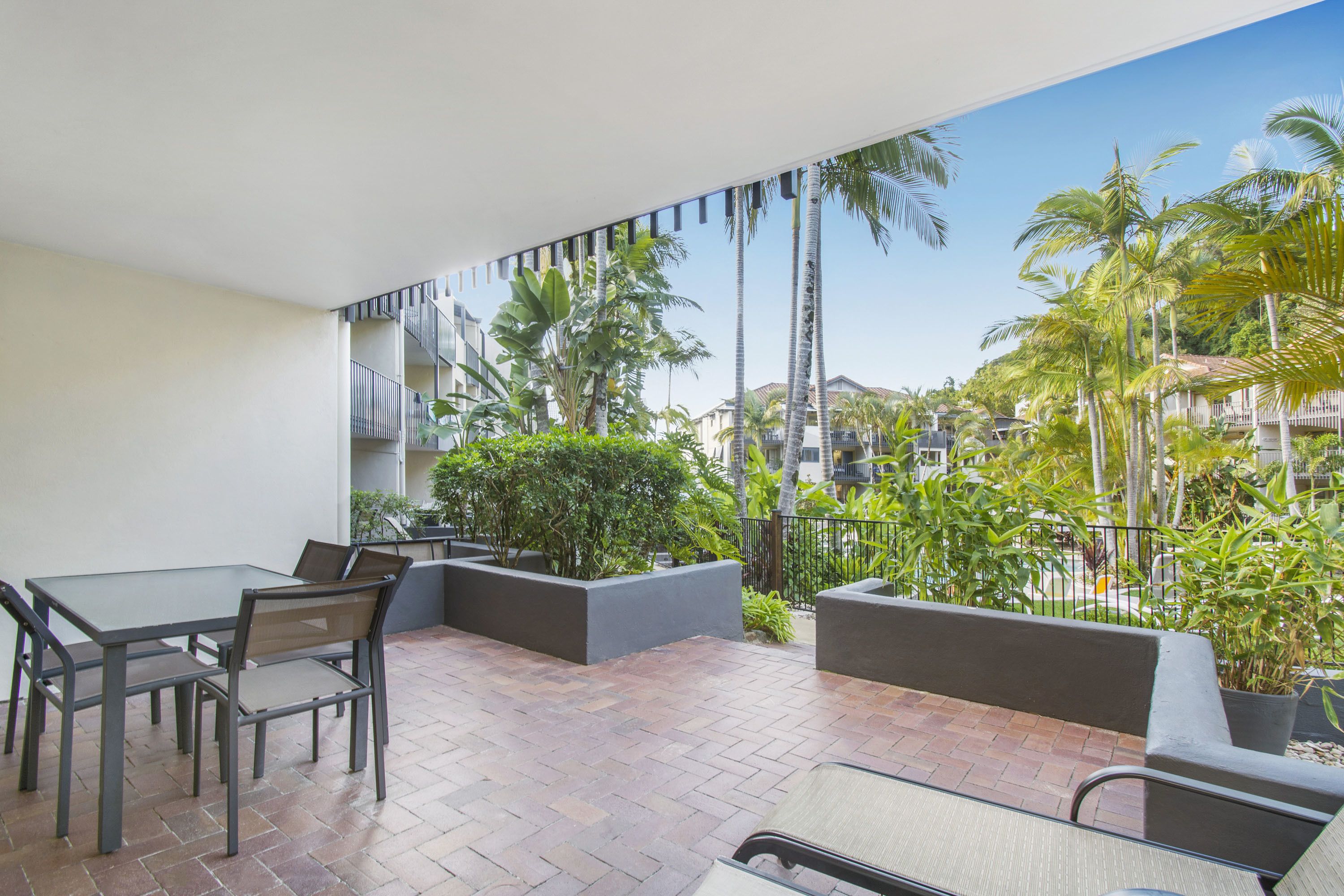 Un balcon avec une table et des chaises, entouré de plantes en pot et offrant une vue sur des palmiers et des immeubles.