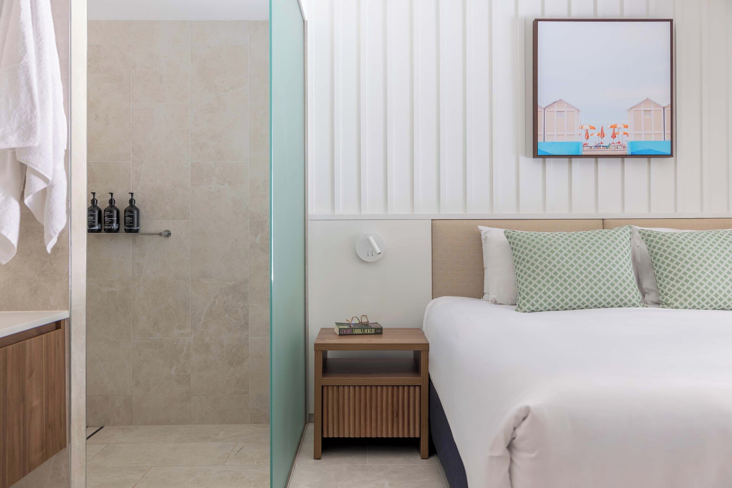 Een hotelkamer met een bed, een nachtkastje, een klein plankje met toiletartikelen en een ingelijst schilderij aan de muur.