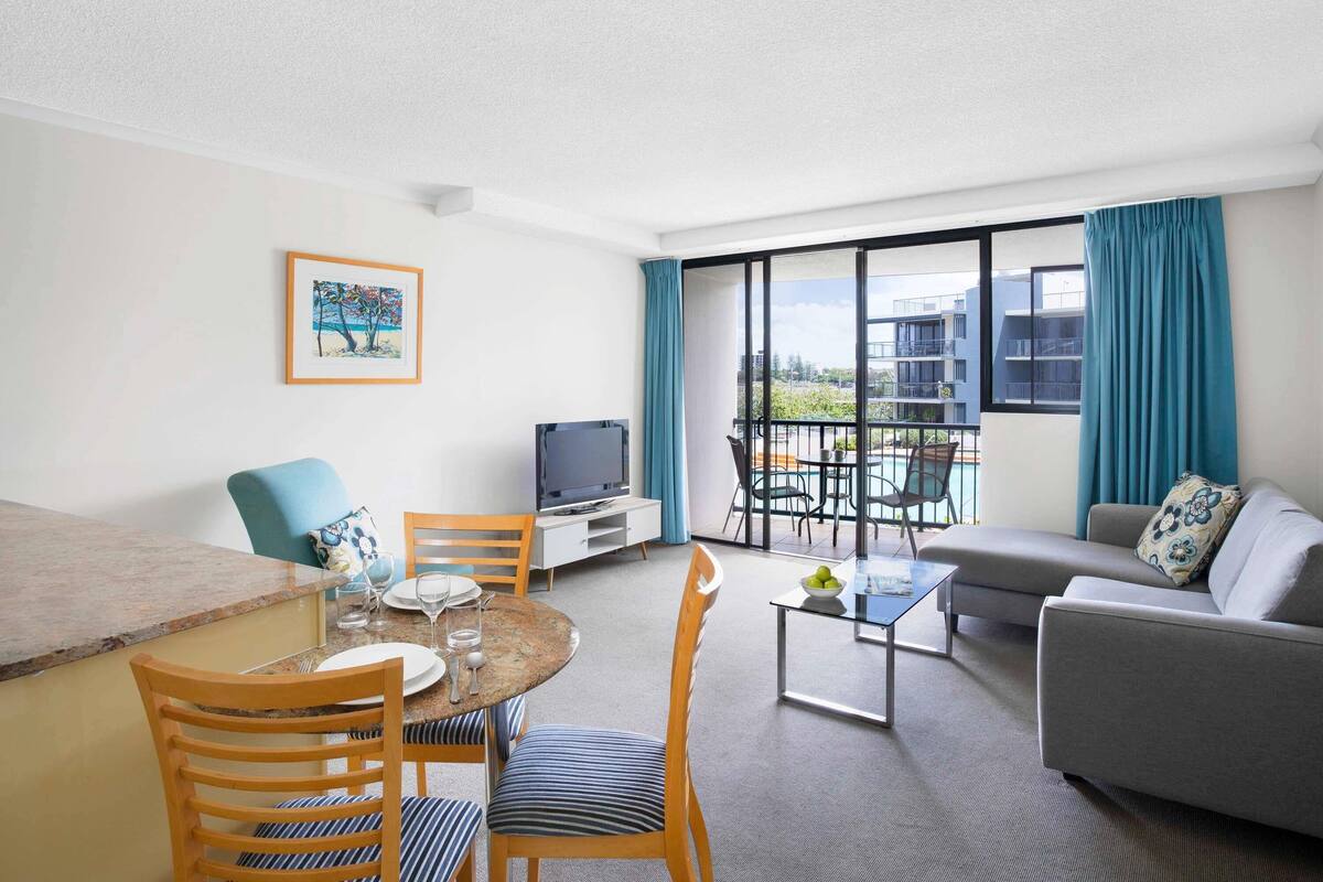 A hotel room with a dining area, a TV, a sofa, and a balcony with a view.