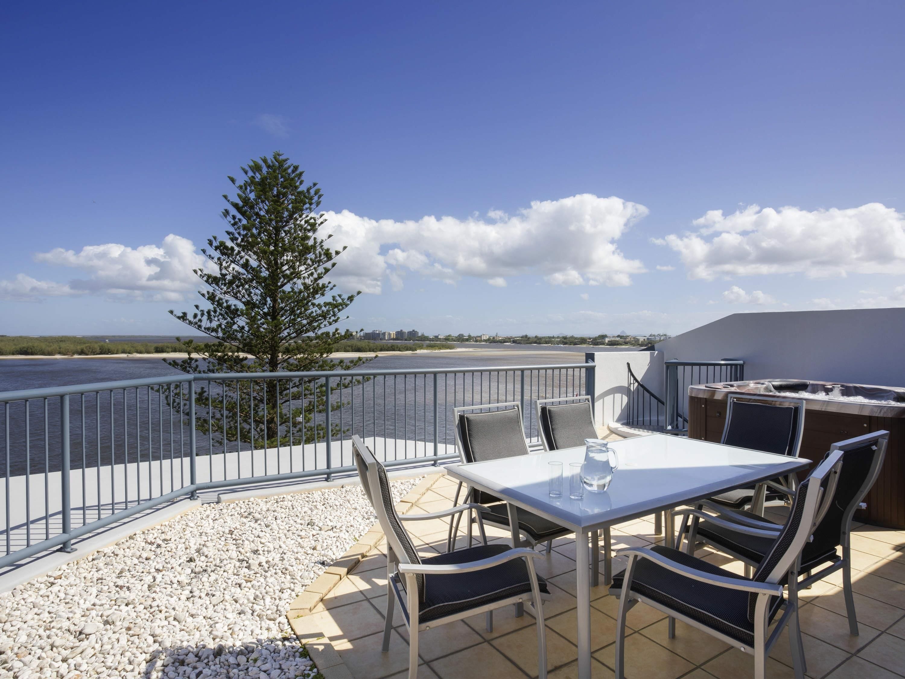 A balcony with a table and chairs, overlooking a body of water and a distant shoreline.