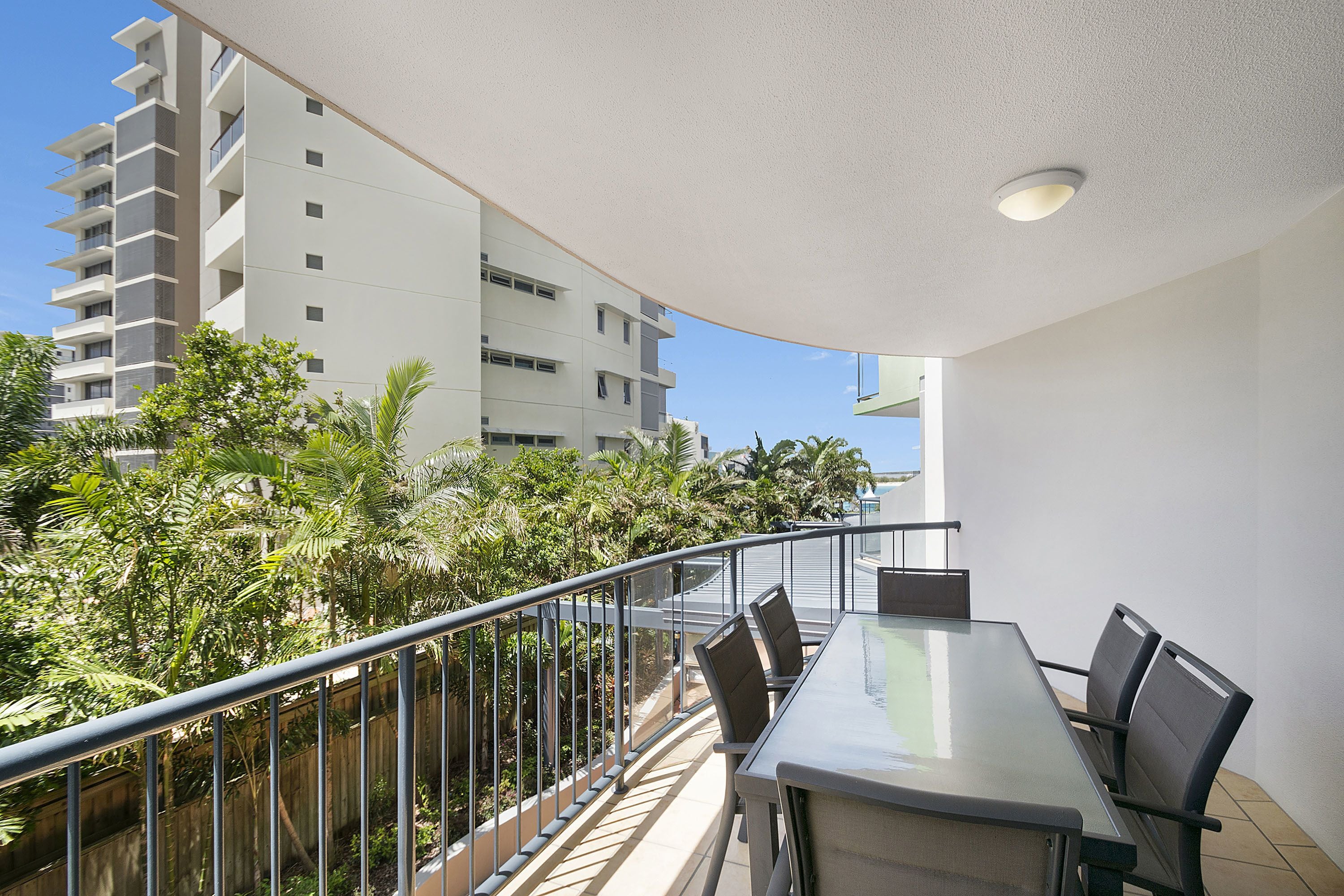 A balcony with a glass table and chairs, overlooking a building and lush greenery.