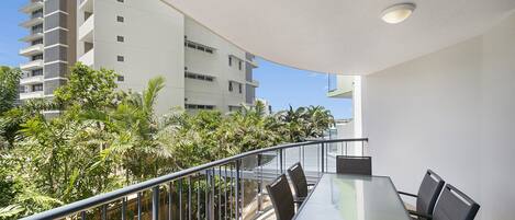 A balcony with a glass table and chairs, overlooking a building and lush greenery.