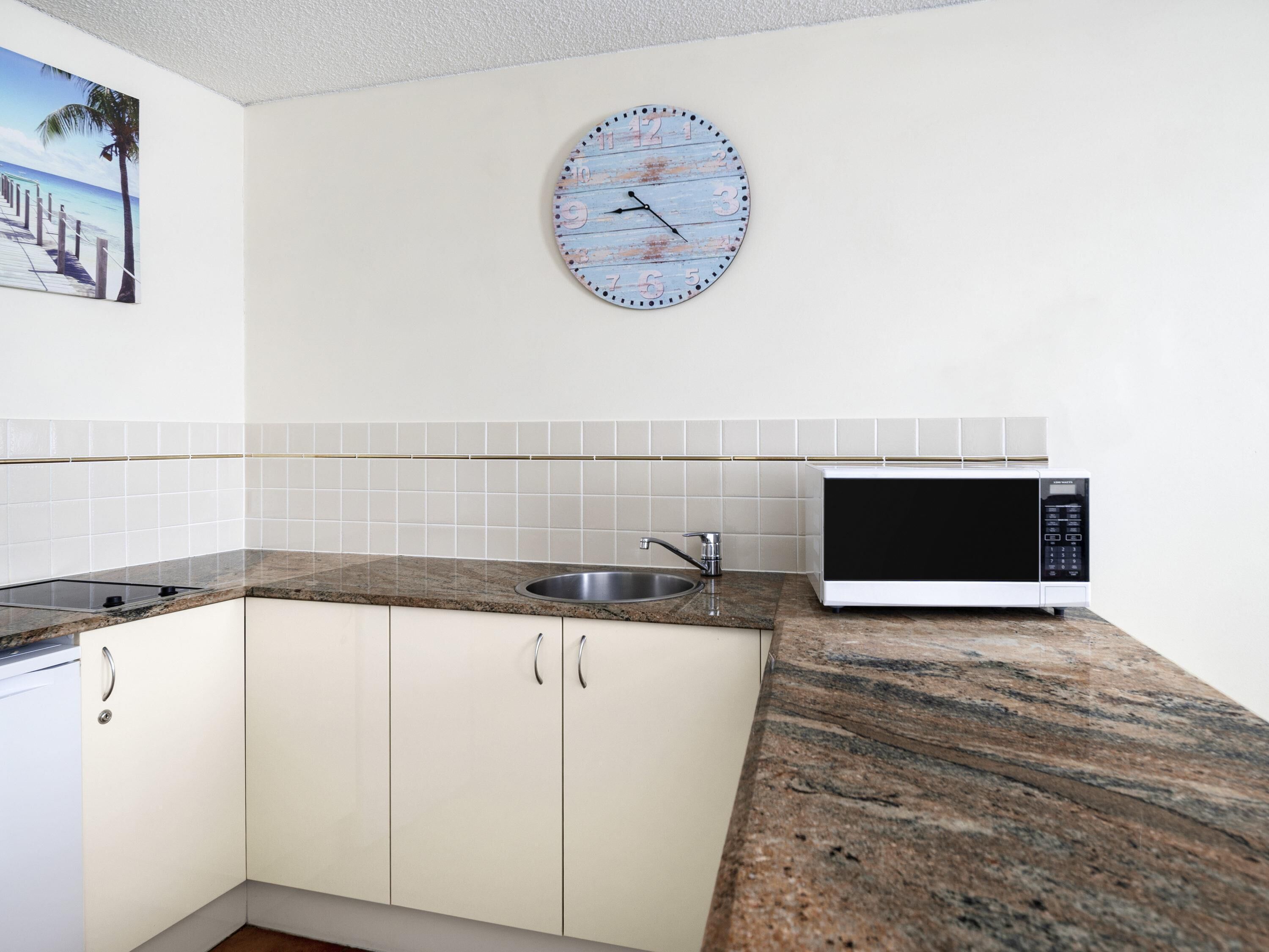 Una cocina con armarios blancos, encimera de granito, pileta de acero inoxidable, microondas blanco y un reloj con una escena de playa.