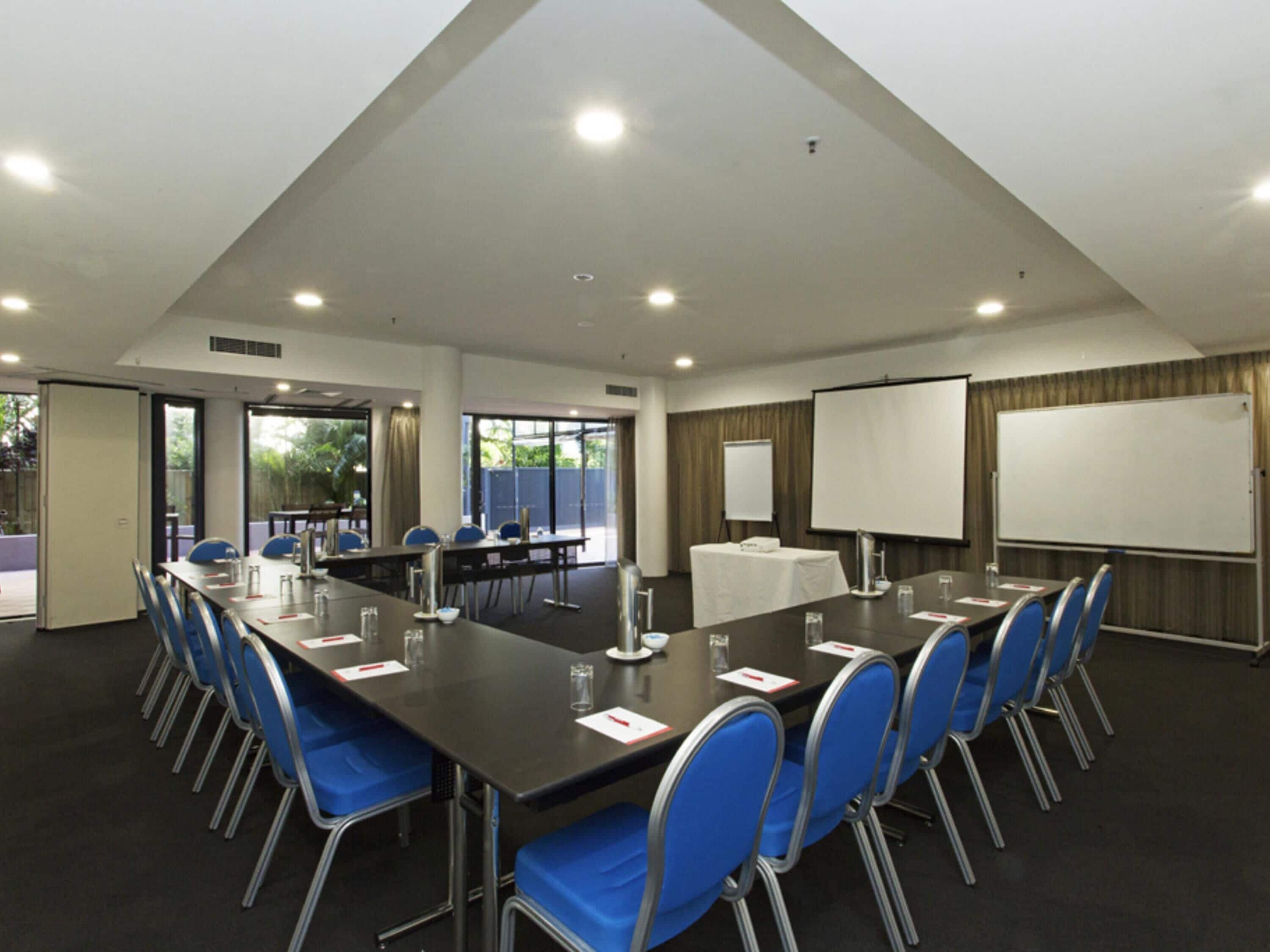 Uma sala de conferências com uma mesa comprida, cadeiras azuis, um quadro branco e uma tela de projetor.