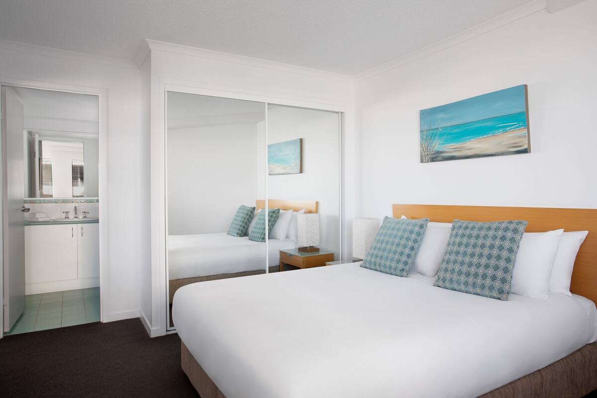 A hotel room with a large bed, a bathroom with a sink and cabinets, and a painting of a beach scene on the wall.