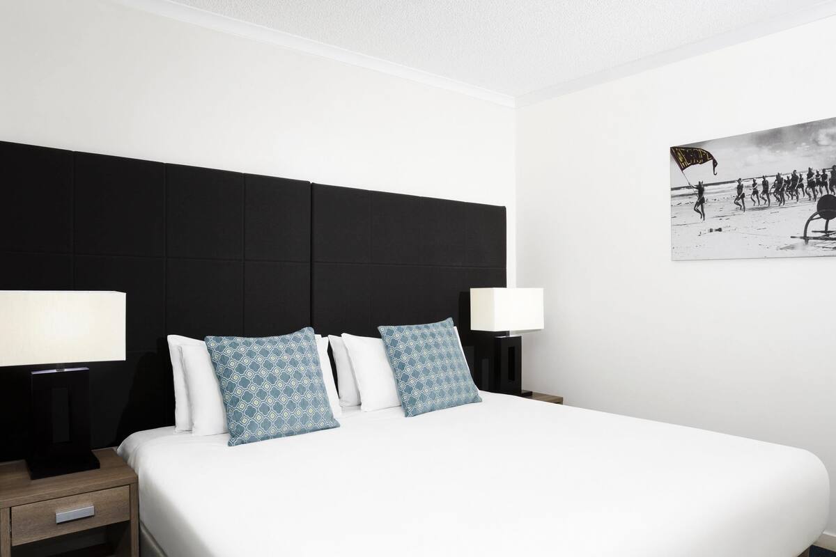 A hotel room with a large bed, two bedside tables with lamps, a black headboard, and a wall-mounted picture.