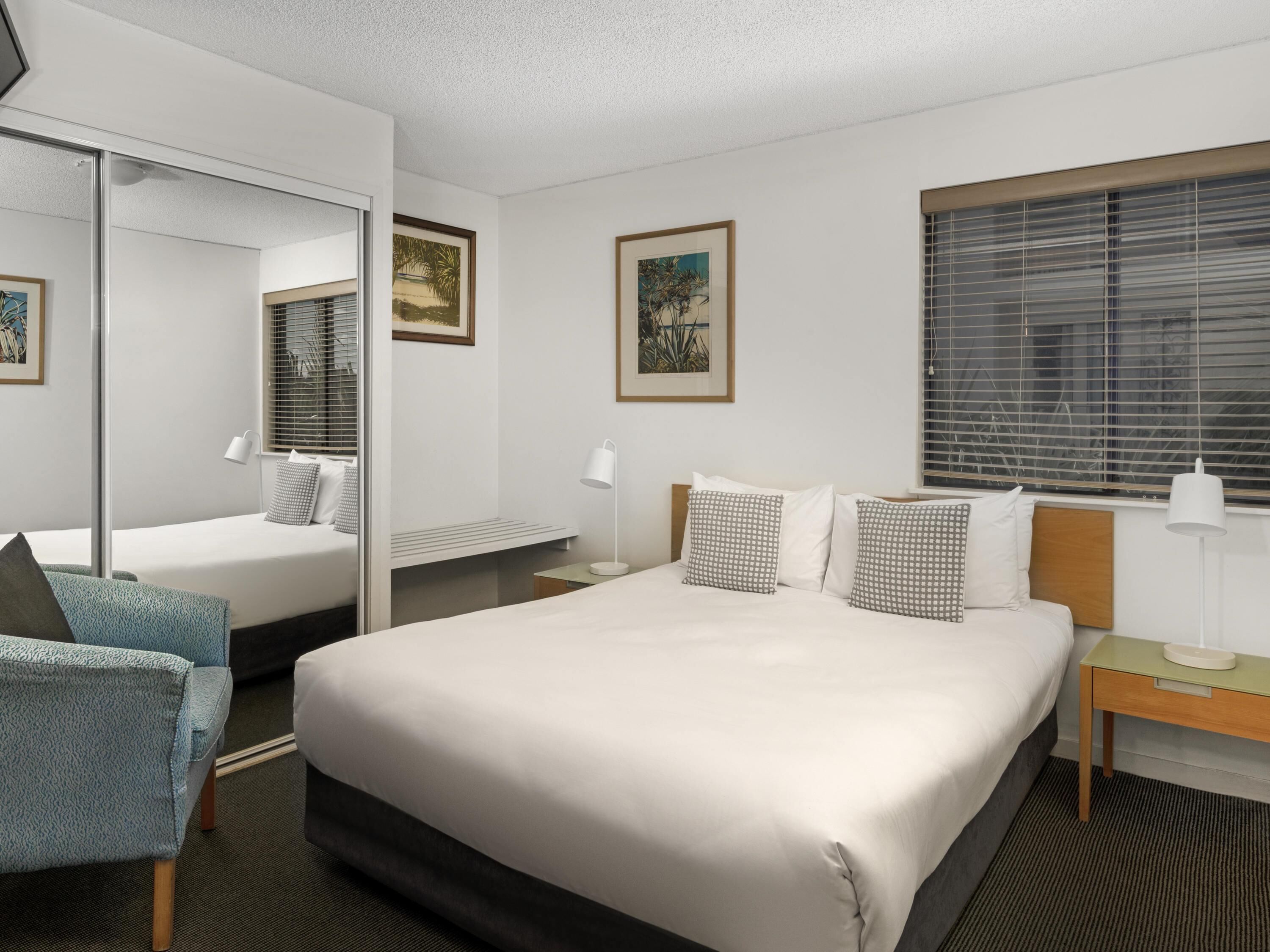 A hotel room with a large bed, a desk, a chair, a mirror, and two windows with blinds.