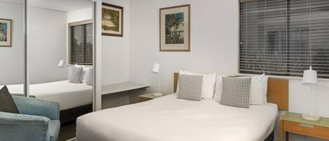A hotel room with a large bed, a desk, a chair, a mirror, and two windows with blinds.