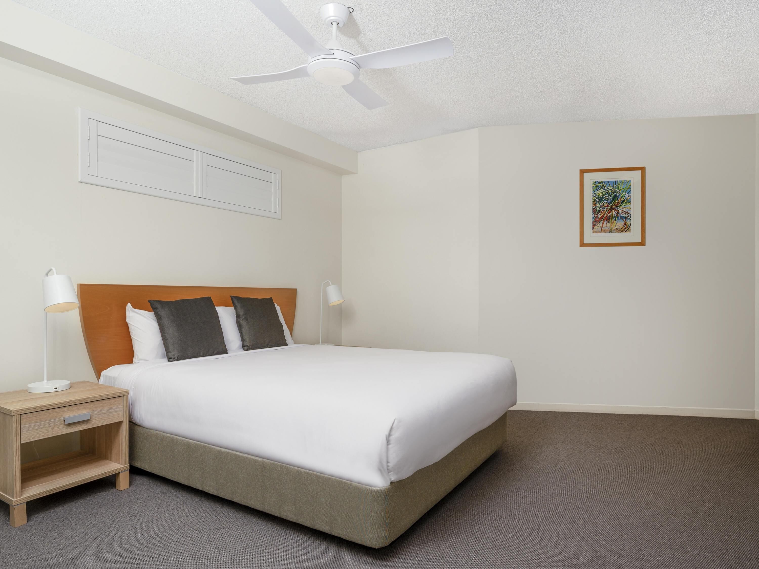 A hotel room with a bed, a nightstand, a ceiling fan, a framed picture on the wall, and a carpeted floor.