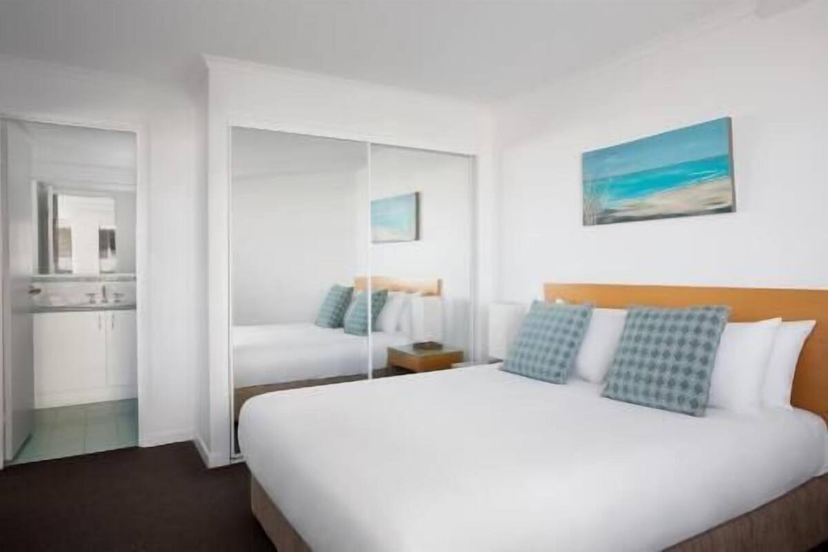 A hotel room with a large bed, a nightstand, a mirror, a painting of a beach, and a bathroom with a sink and mirror.