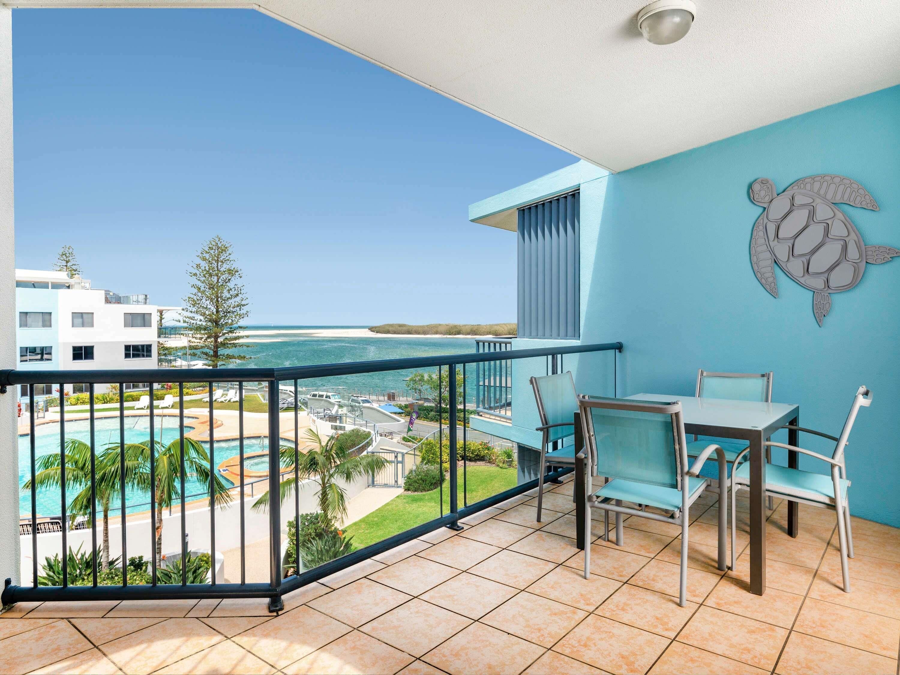 A balcony with a view of a pool and beach, a table with chairs, and a turtle mural on the wall.