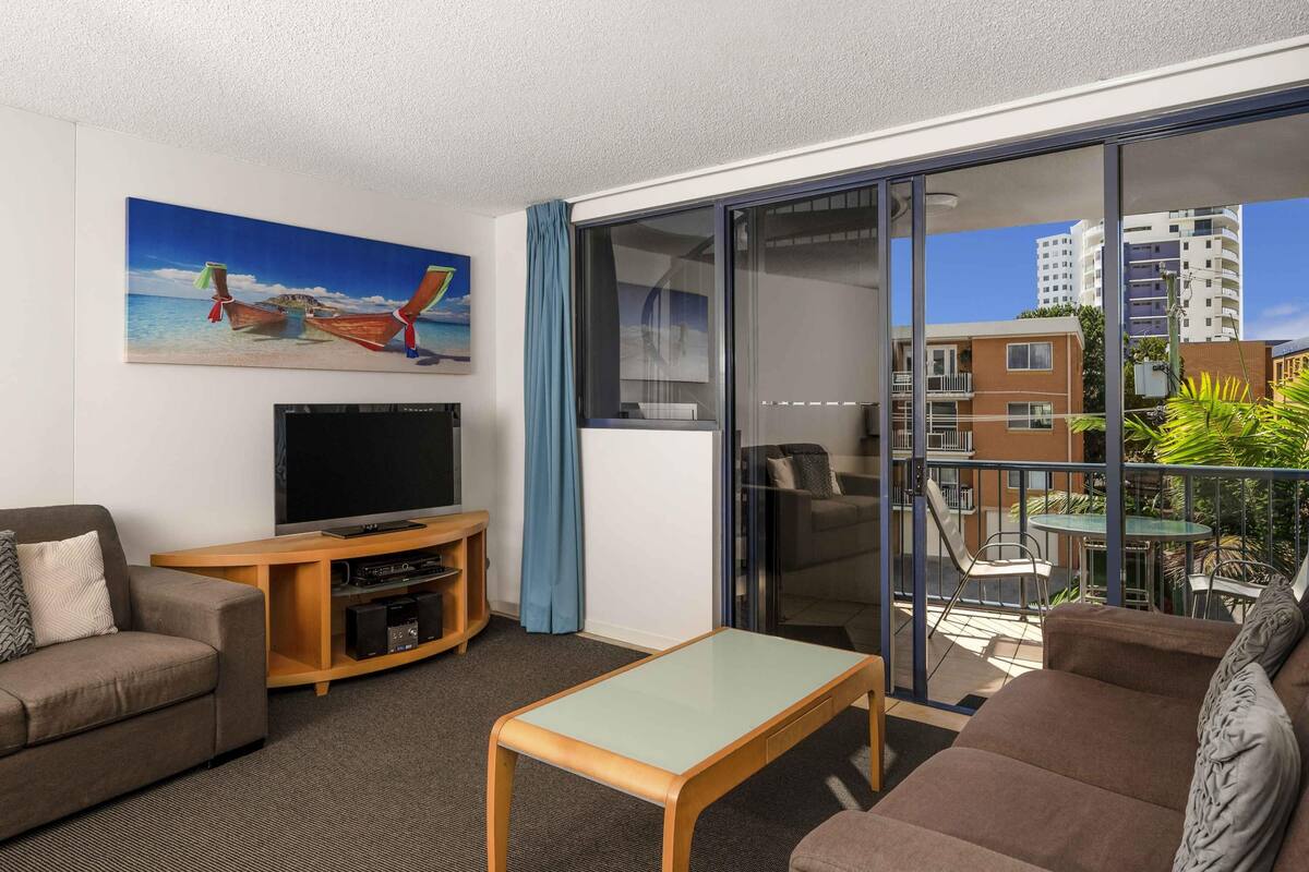 A modern living room with a flat-screen TV, a sofa, a coffee table, and a painting of a boat on a beach.