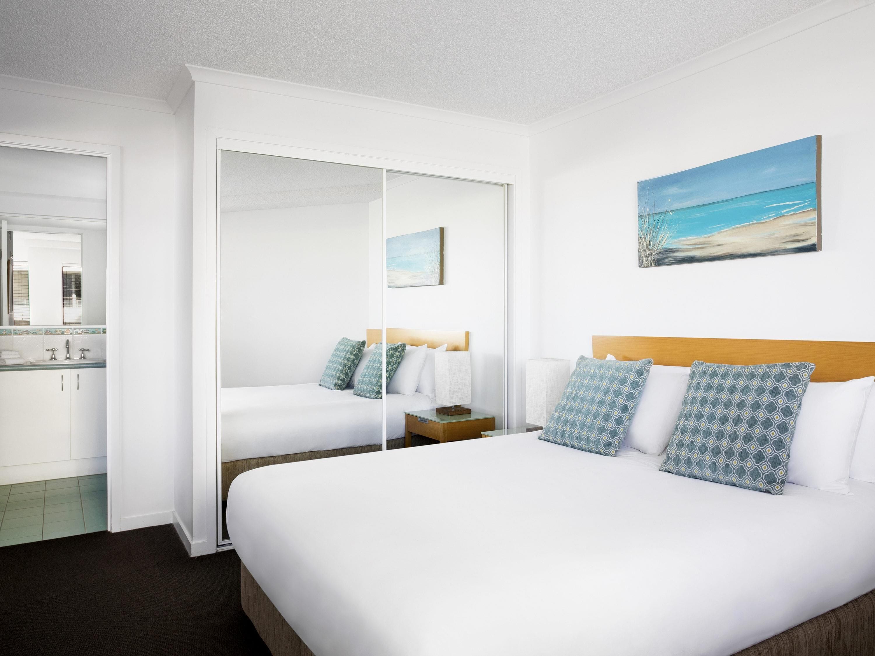 A hotel room with a bed, a nightstand, a mirror, a painting of a beach, and a bathroom with a sink and toilet.