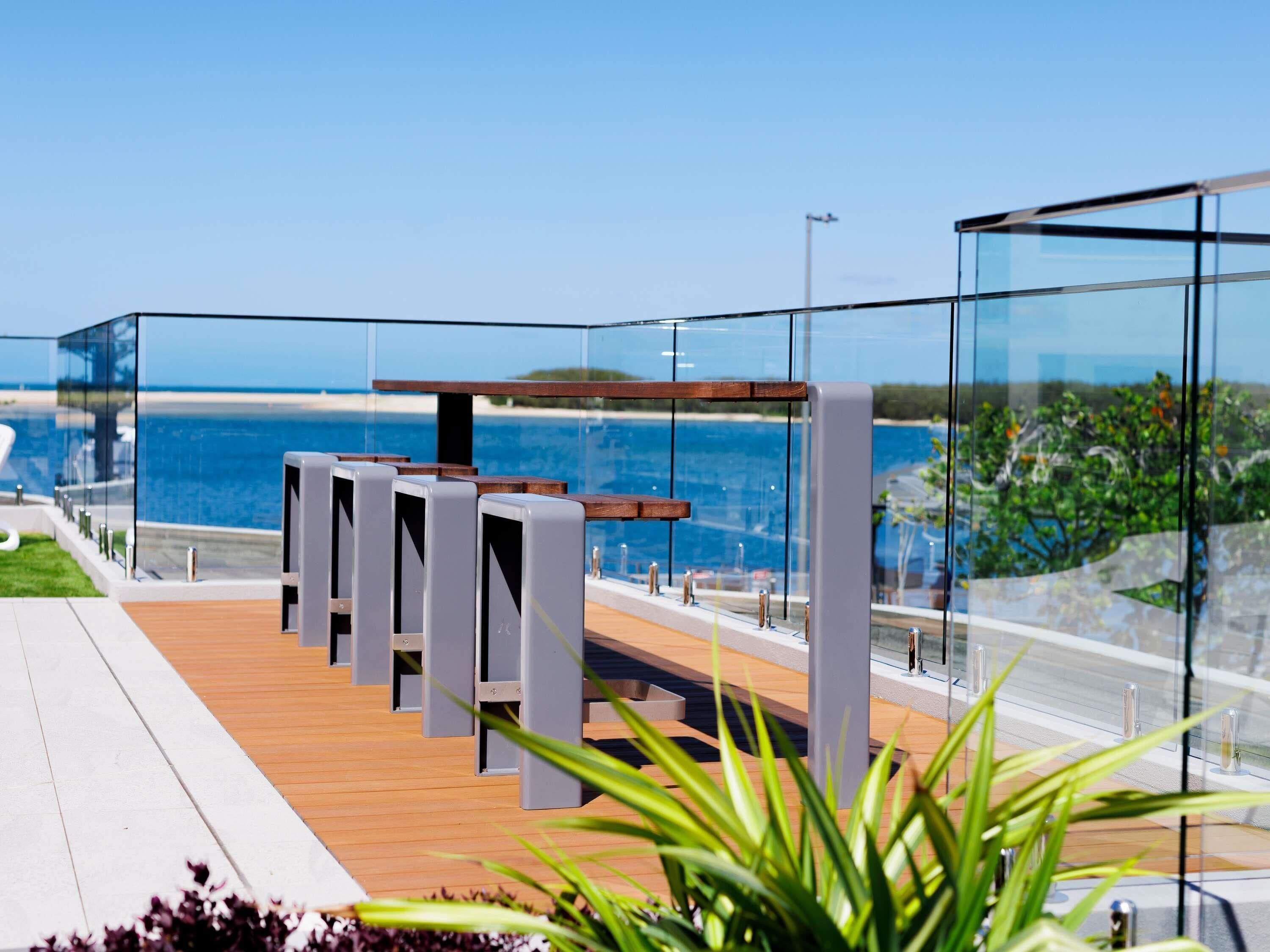 A modern outdoor seating area with glass railings, wooden benches, and a view of the sea.
